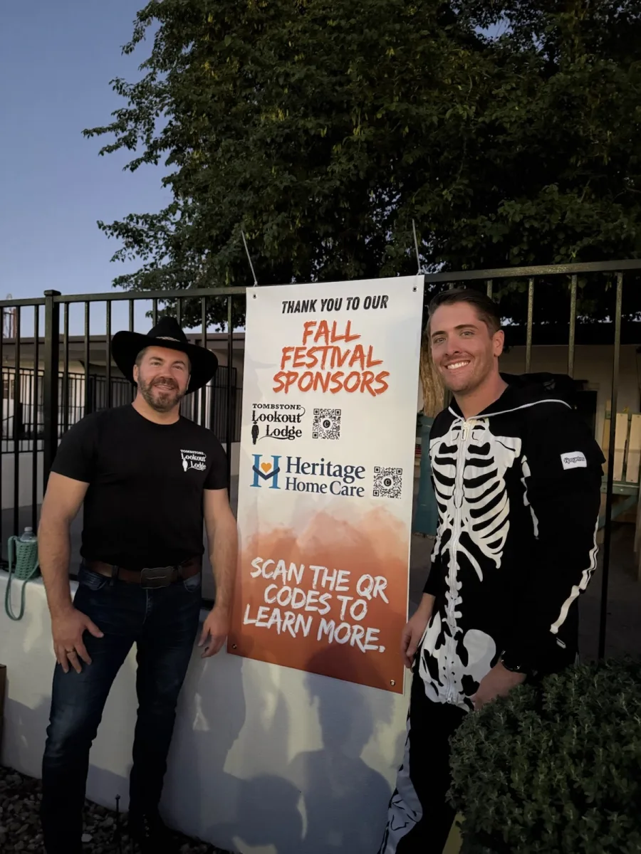 Two festival participants standing beside a Fall Festival sponsor sign featuring Heritage Home Care.