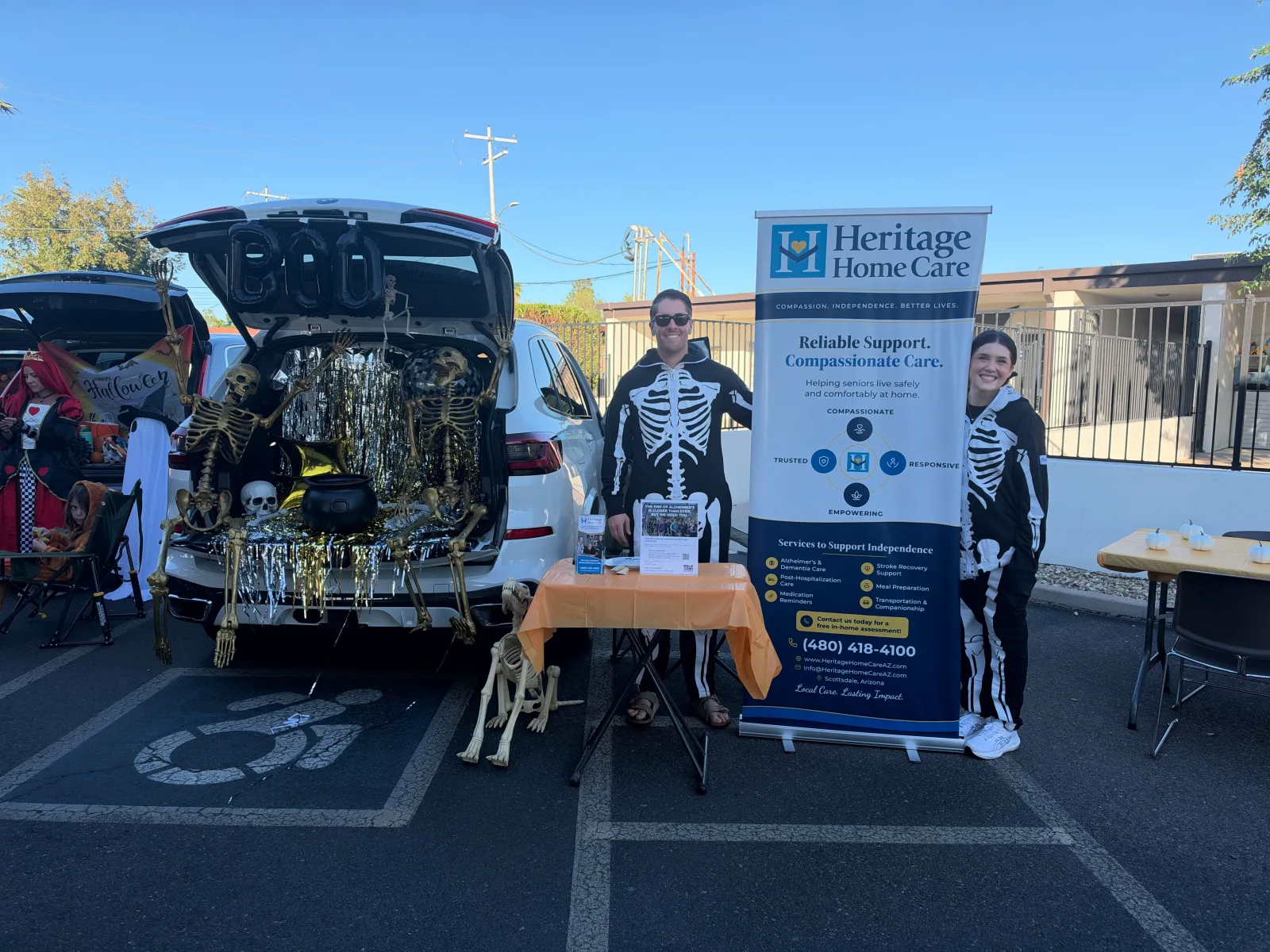 The Heritage team in skeleton costumes beside their decorated Trunk or Treat vehicle and Heritage Home Care banner.