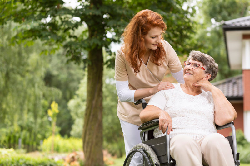 Caregiver walking alongside an elderly woman in a sunny garden, both smiling